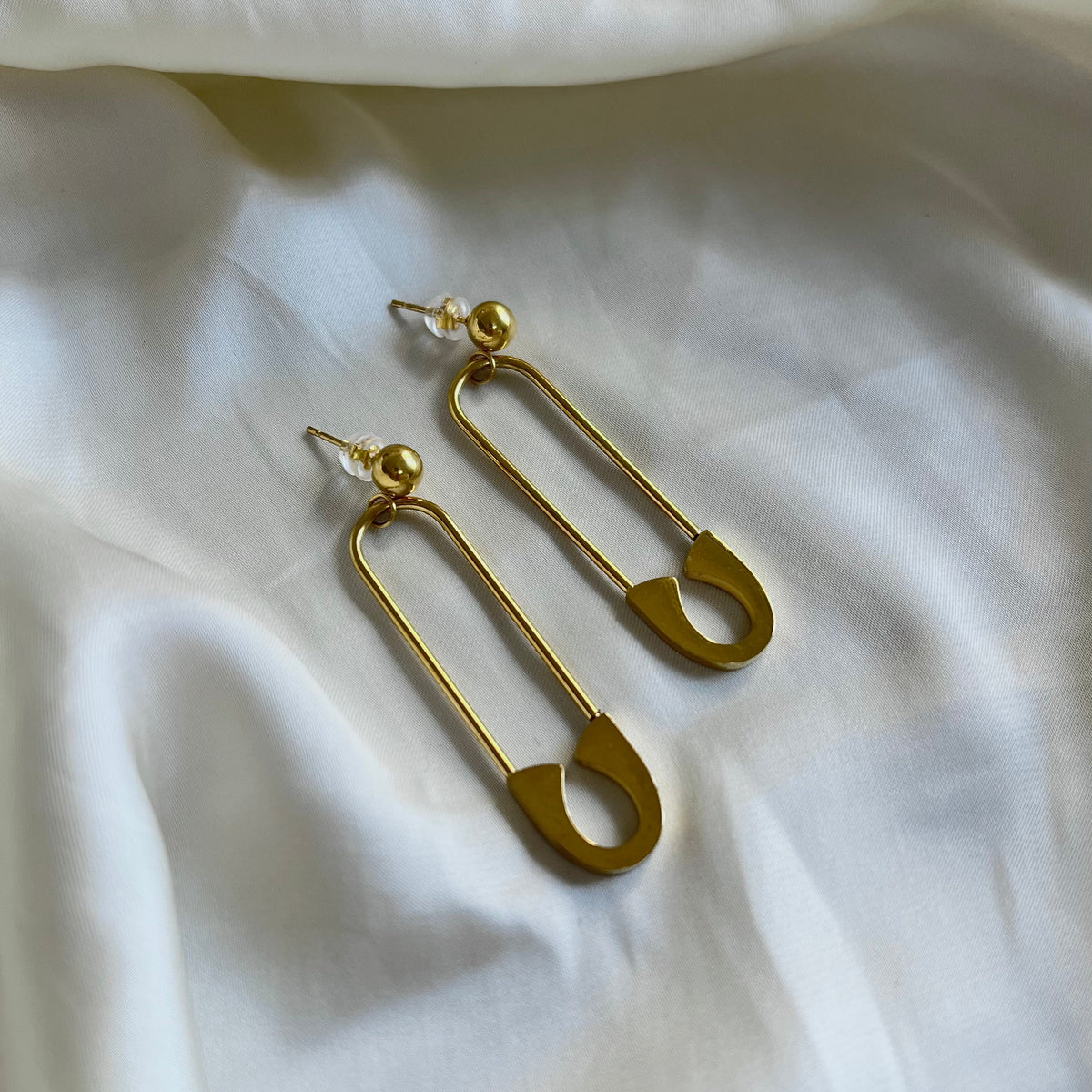 A pair of gold-plated safety pin drop earrings hanging from round studs on ivory fabric